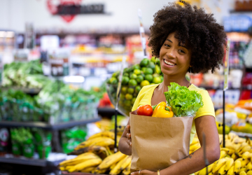 A bright and smiling young woman holding a full bag of fresh produce and groceries.